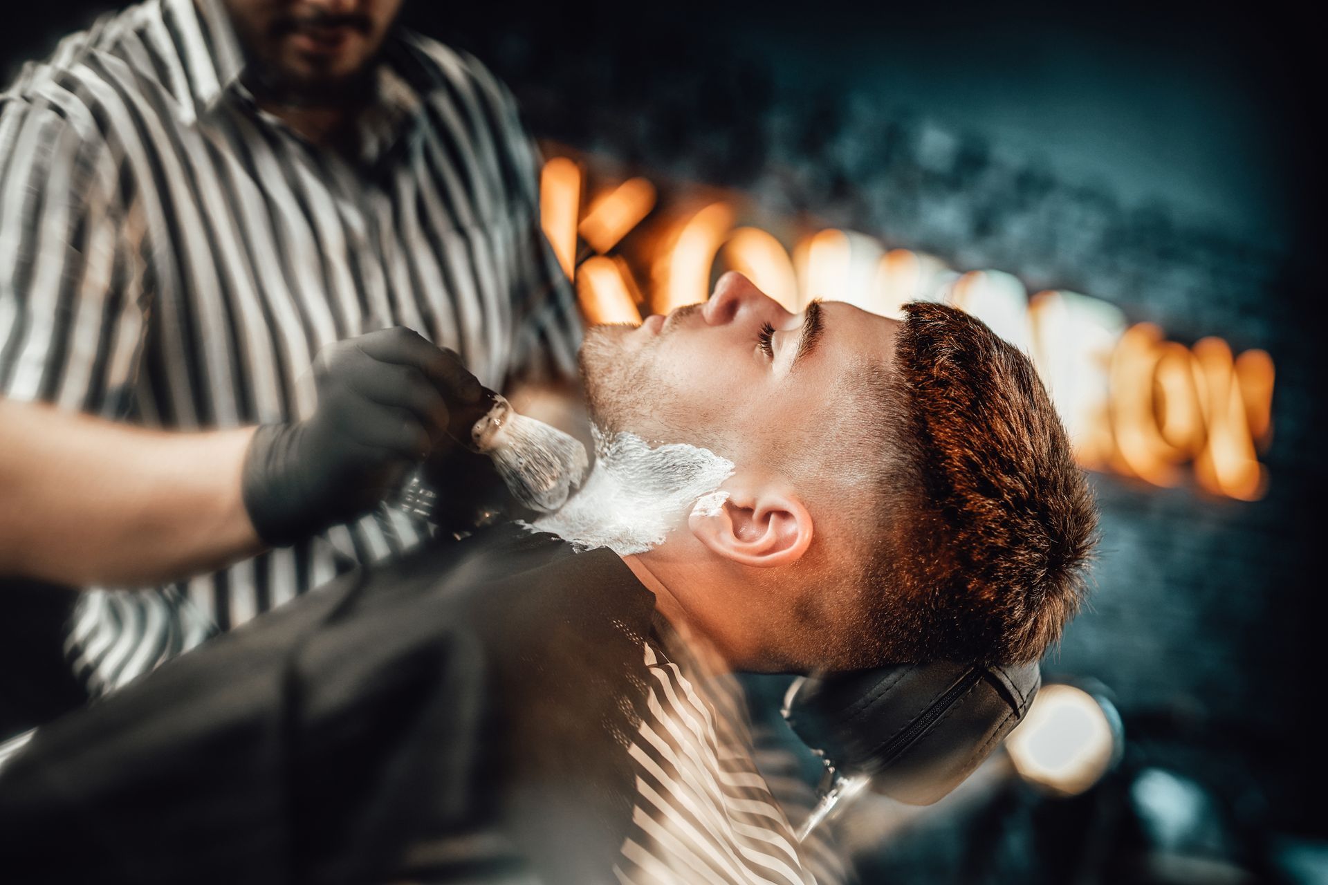 Dans un salon de coiffure, un barbier en chemise rayée applique de la mousse à raser sur le visage d'un client.