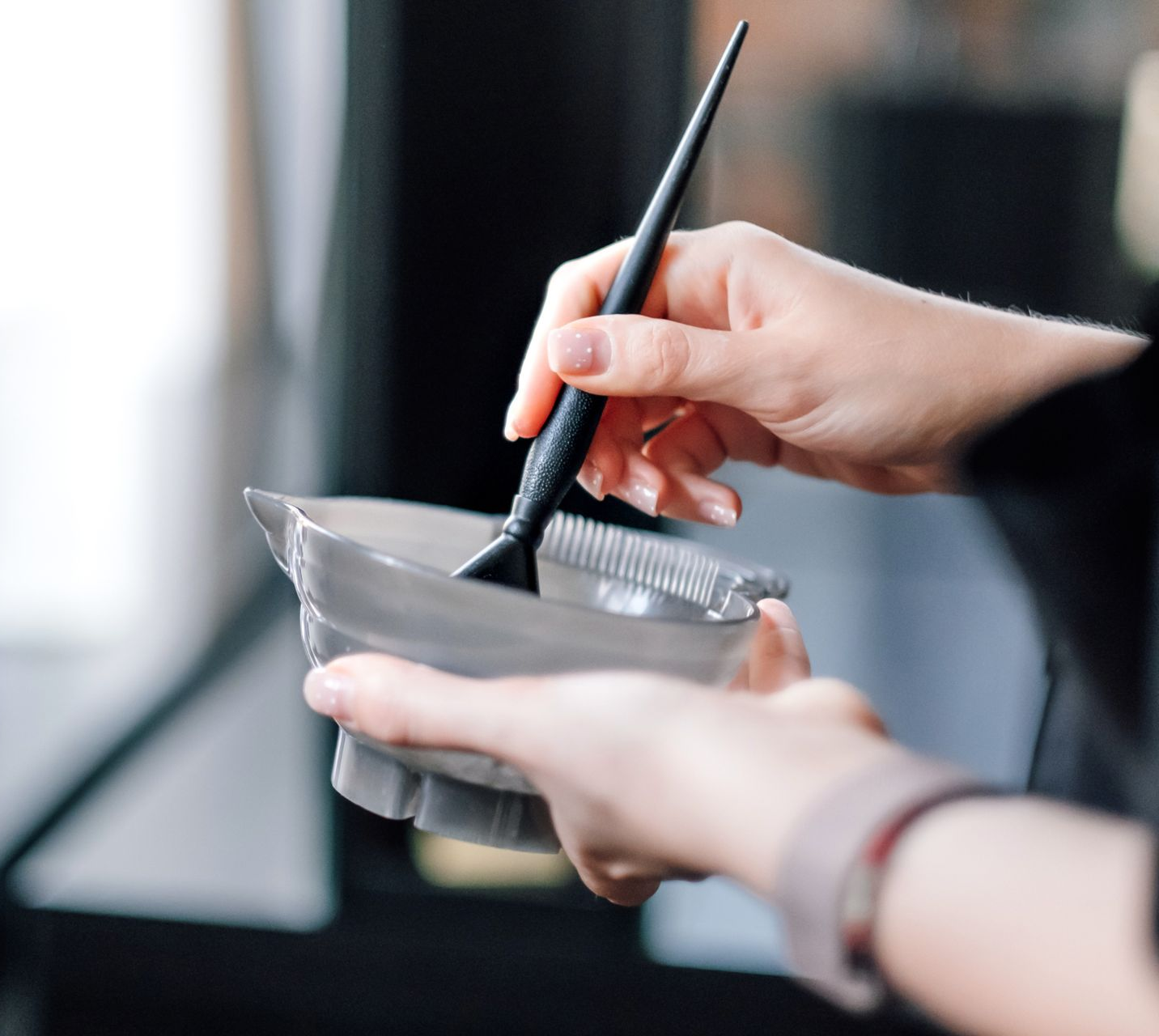 Gros plan sur des mains tenant un bol mélangeur gris et un pinceau pour teinture capillaire.
