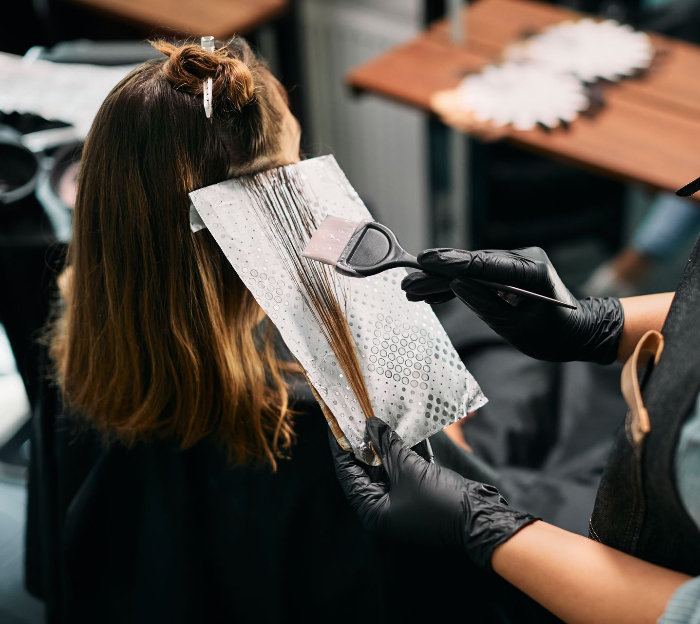 Une coiffeuse portant des gants noirs utilise un pinceau pour appliquer une coloration sur une mèche de cheveux posée sur une feuille d'aluminium.