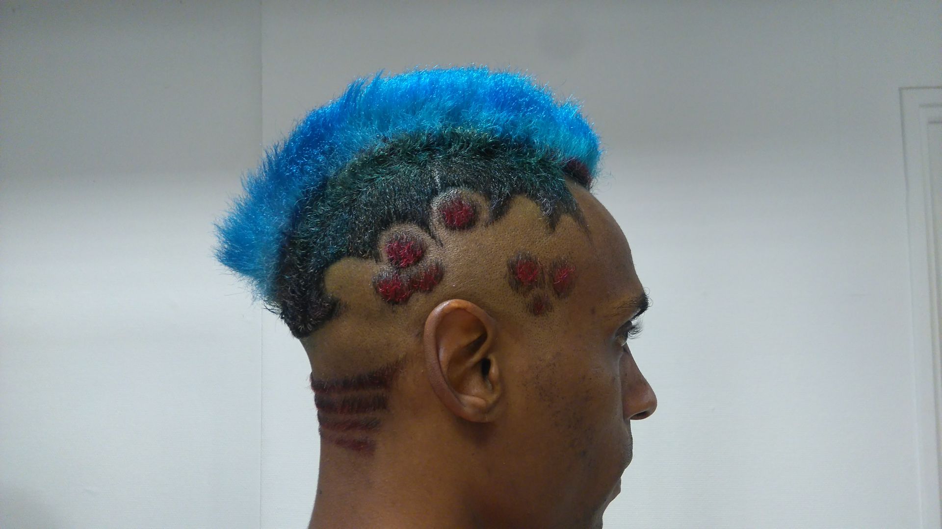 Gros plan sur une personne arborant une crête iroquoise bleu vif, des motifs latéraux sombres à paillettes et des accents circulaires rouges sur la tempe.