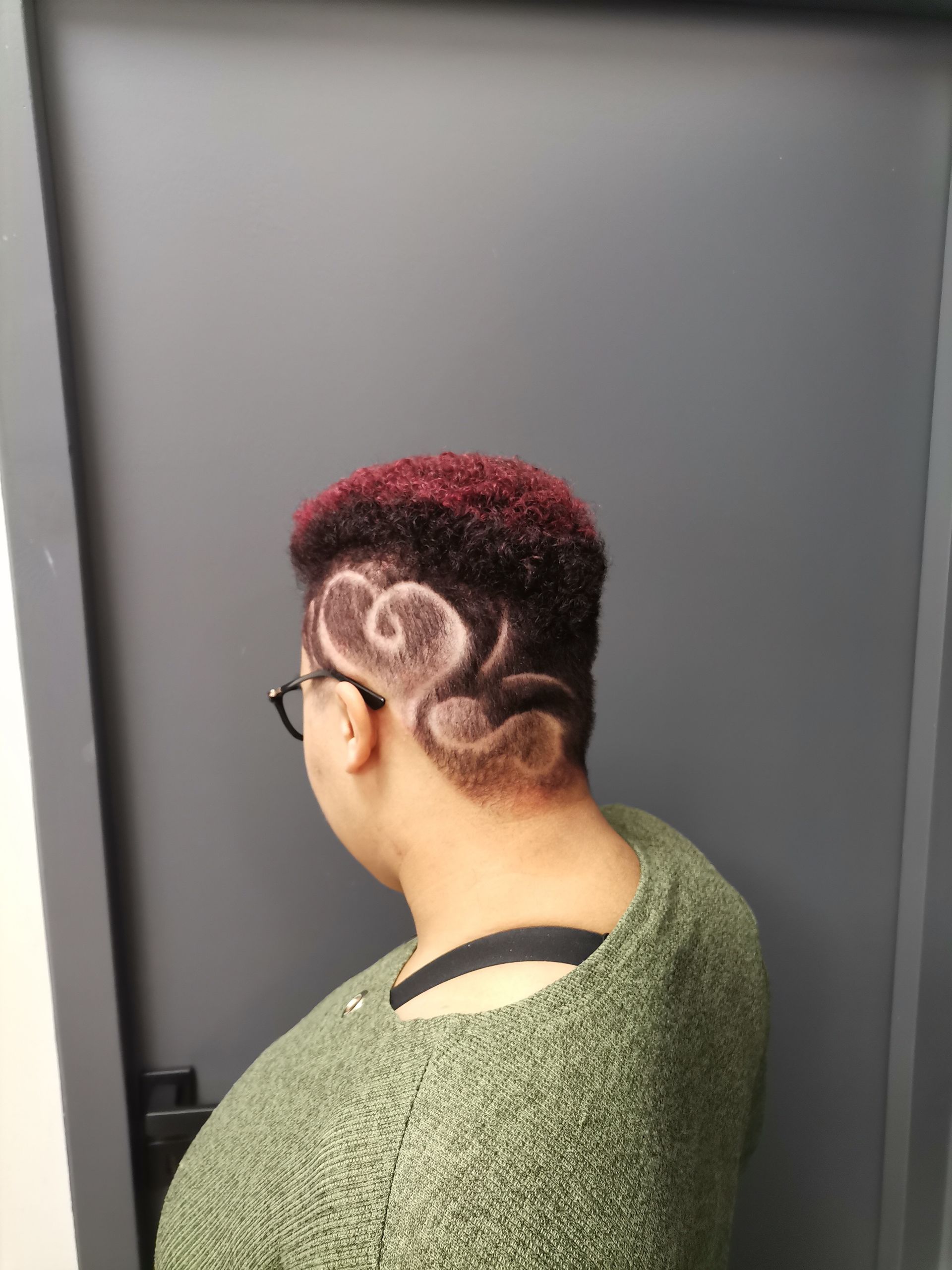 Une personne aux cheveux aux pointes rouge foncé et présentant un motif complexe de spirales rasées sur le côté de la tête.