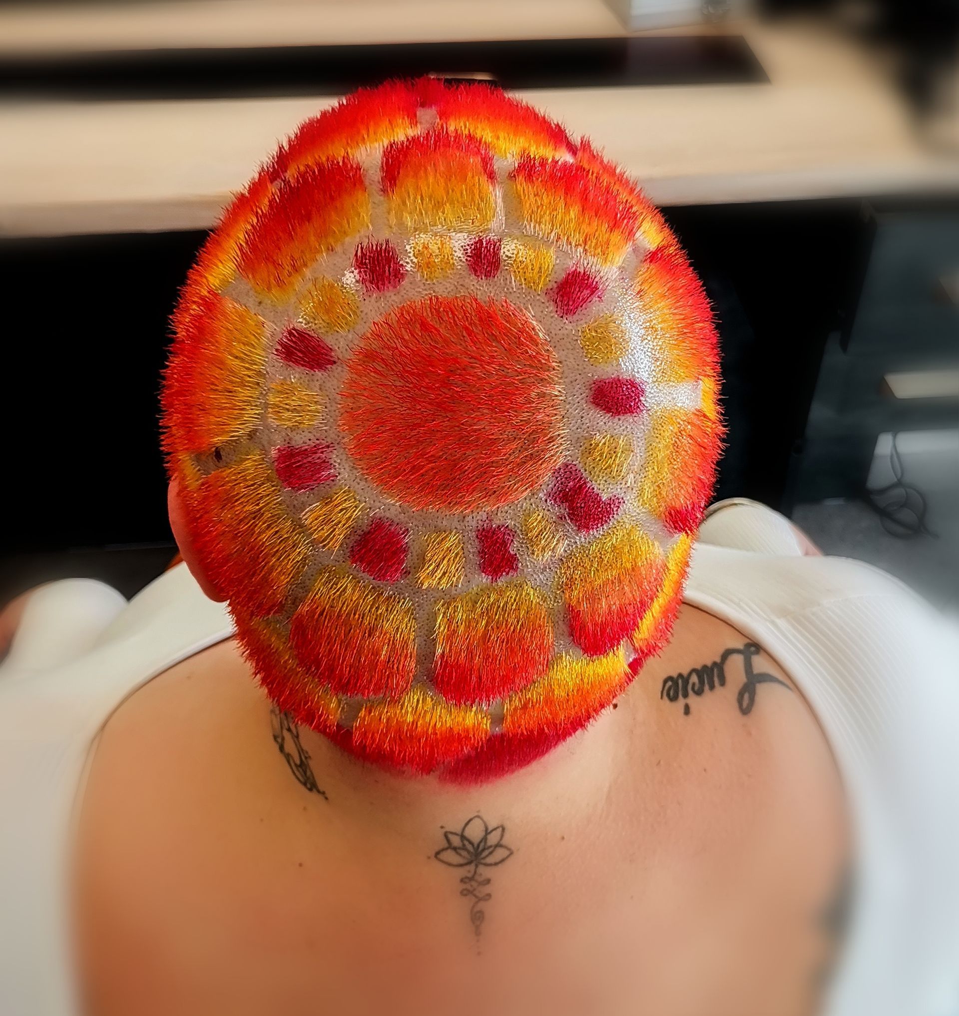 Les cheveux courts d'une personne sont coiffés en un motif circulaire vibrant, inspiré du soleil, avec des dégradés de rouge, d'orange et de jaune.