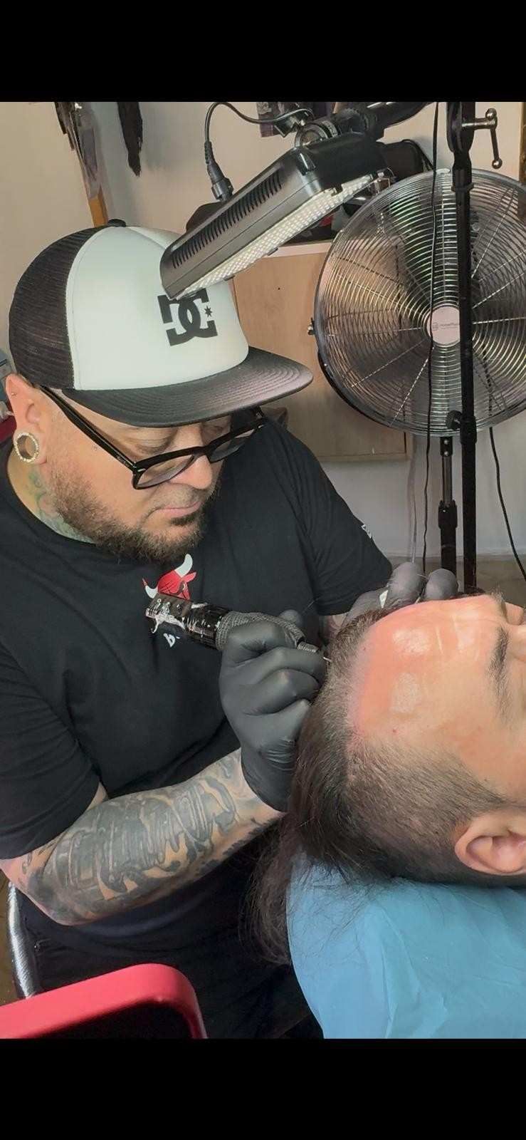 Un tatuador, con guantes y gorro, trabaja en el rostro de un cliente, con un ventilador y un micrófono cerca.