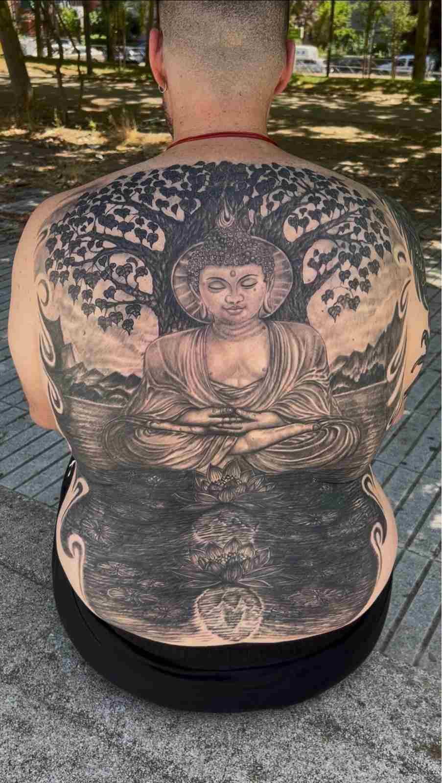 Una persona sentada en un banco muestra un gran tatuaje negro con el diseño de un Buda y un árbol en la espalda.
