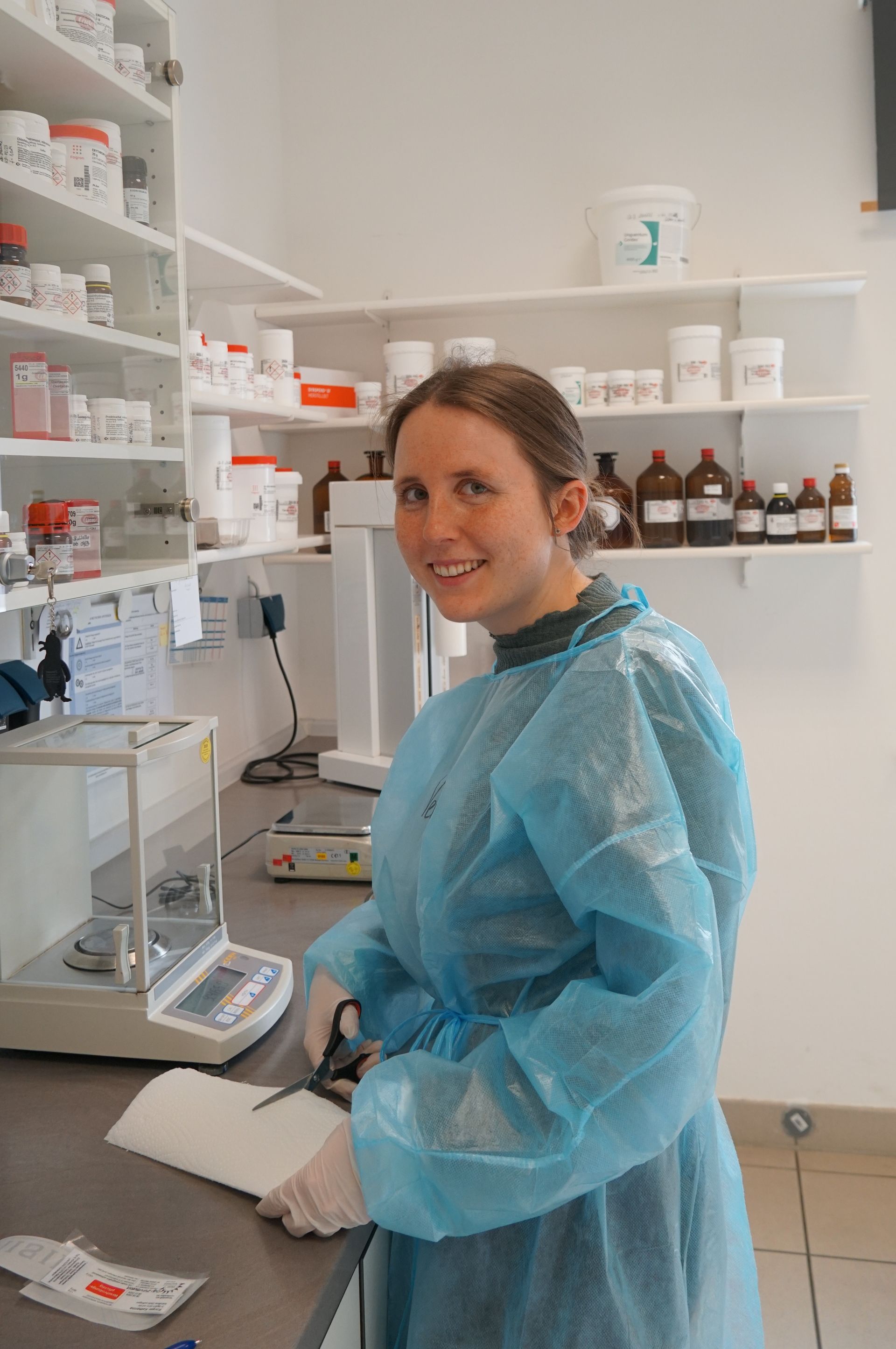 Frau in blauem Kleid schneidet Papier mit einer Schere in einer Apotheke mit Regalen voller Medikamente.