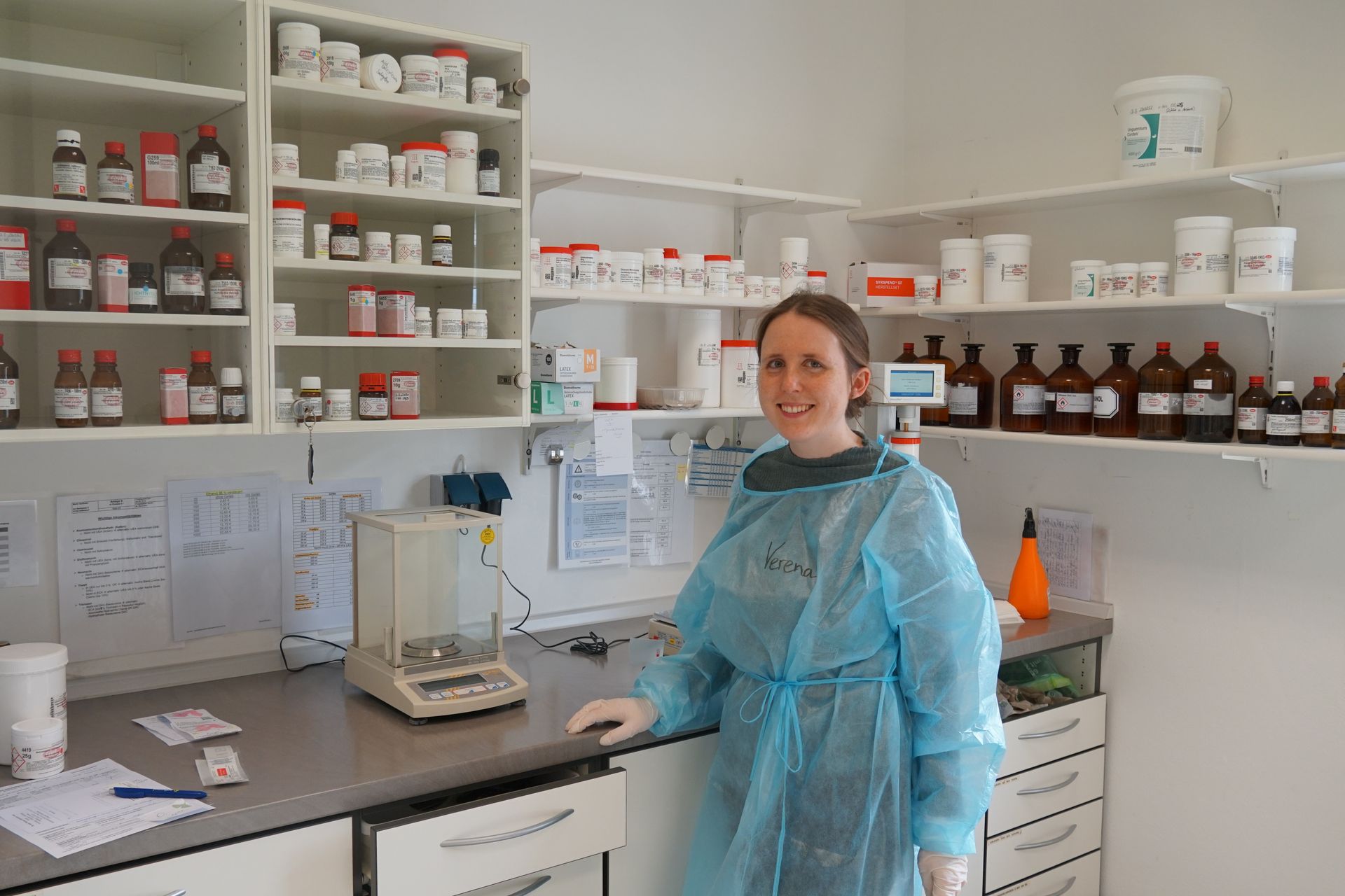 Eine Frau im blauen Laborkittel in einer Apotheke, umgeben von Medikamentenflaschen in Regalen.