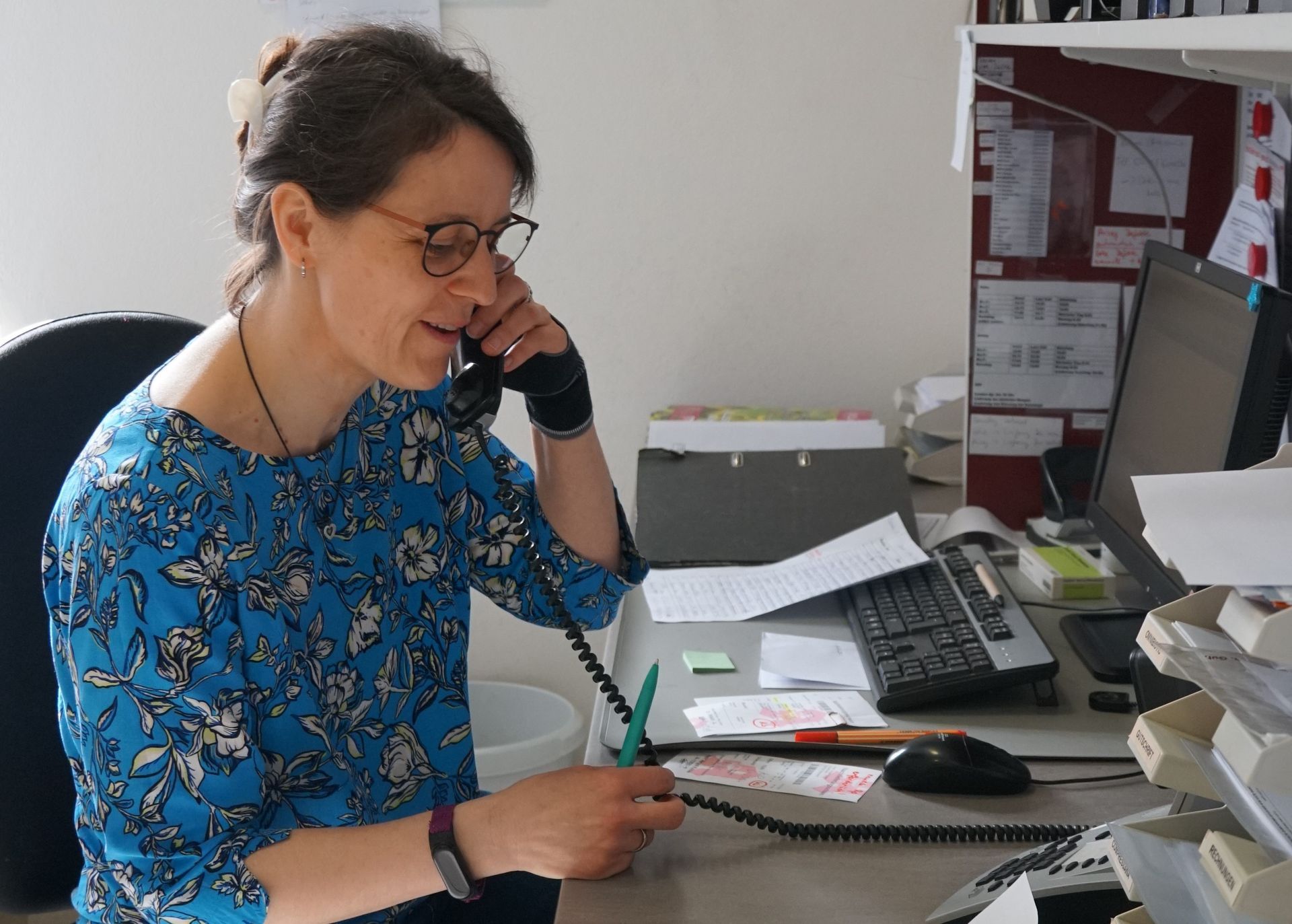 Frau in blauem geblümtem Hemd telefoniert im Büro und blickt auf ihre Unterlagen; Computer und Unterlagen liegen auf dem Schreibtisch.