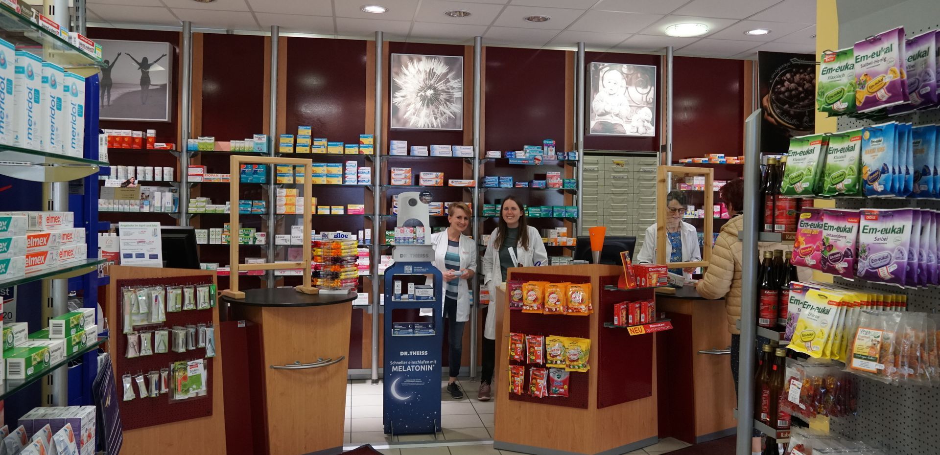 In einer Apotheke stehen drei Personen in weißen Kitteln hinter Theken, an denen Produkte in Regalen ausgestellt sind.