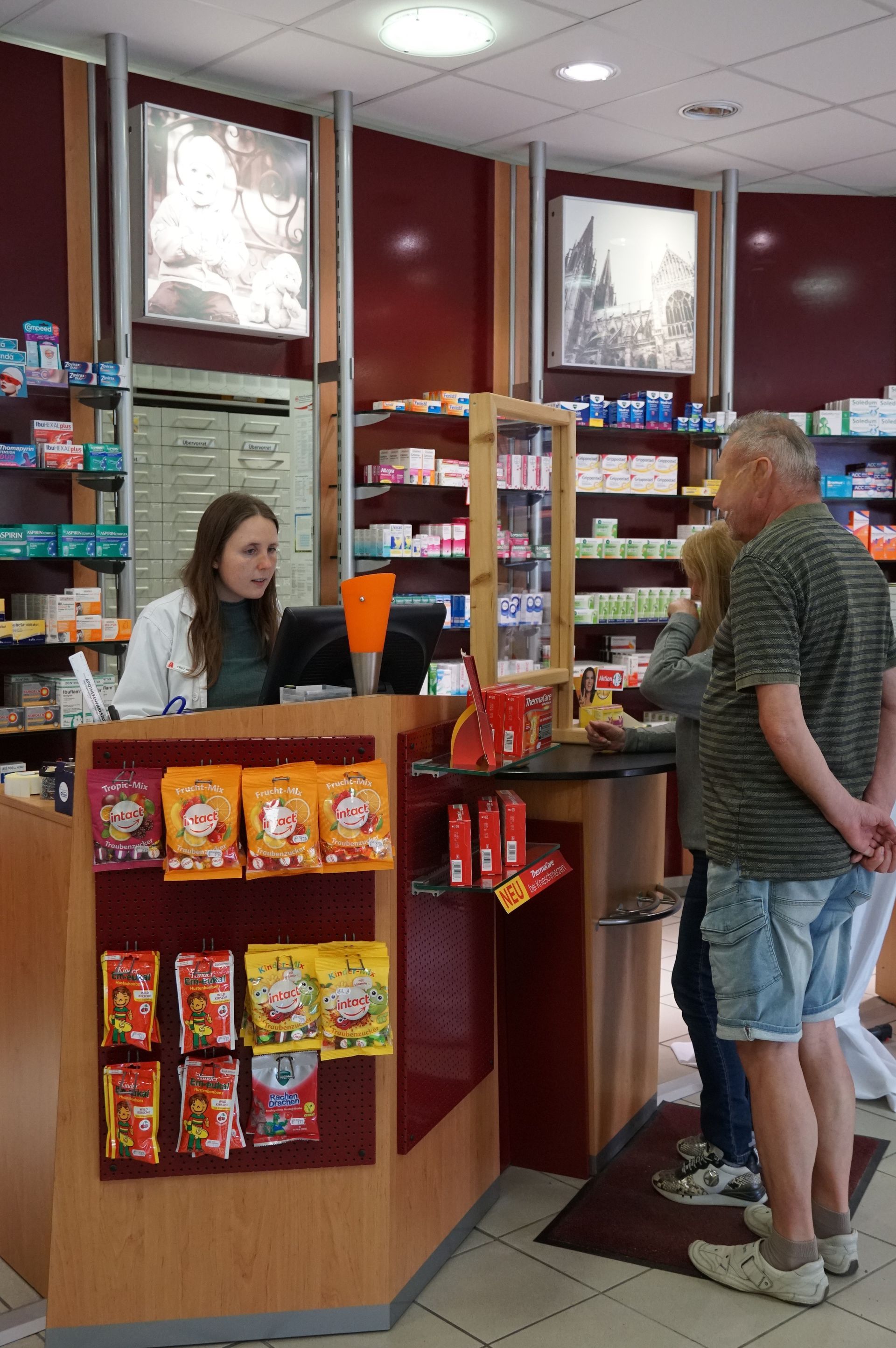 In einer Apotheke bedient ein Apotheker die Kunden am Schalter. In den Regalen werden die Produkte präsentiert.