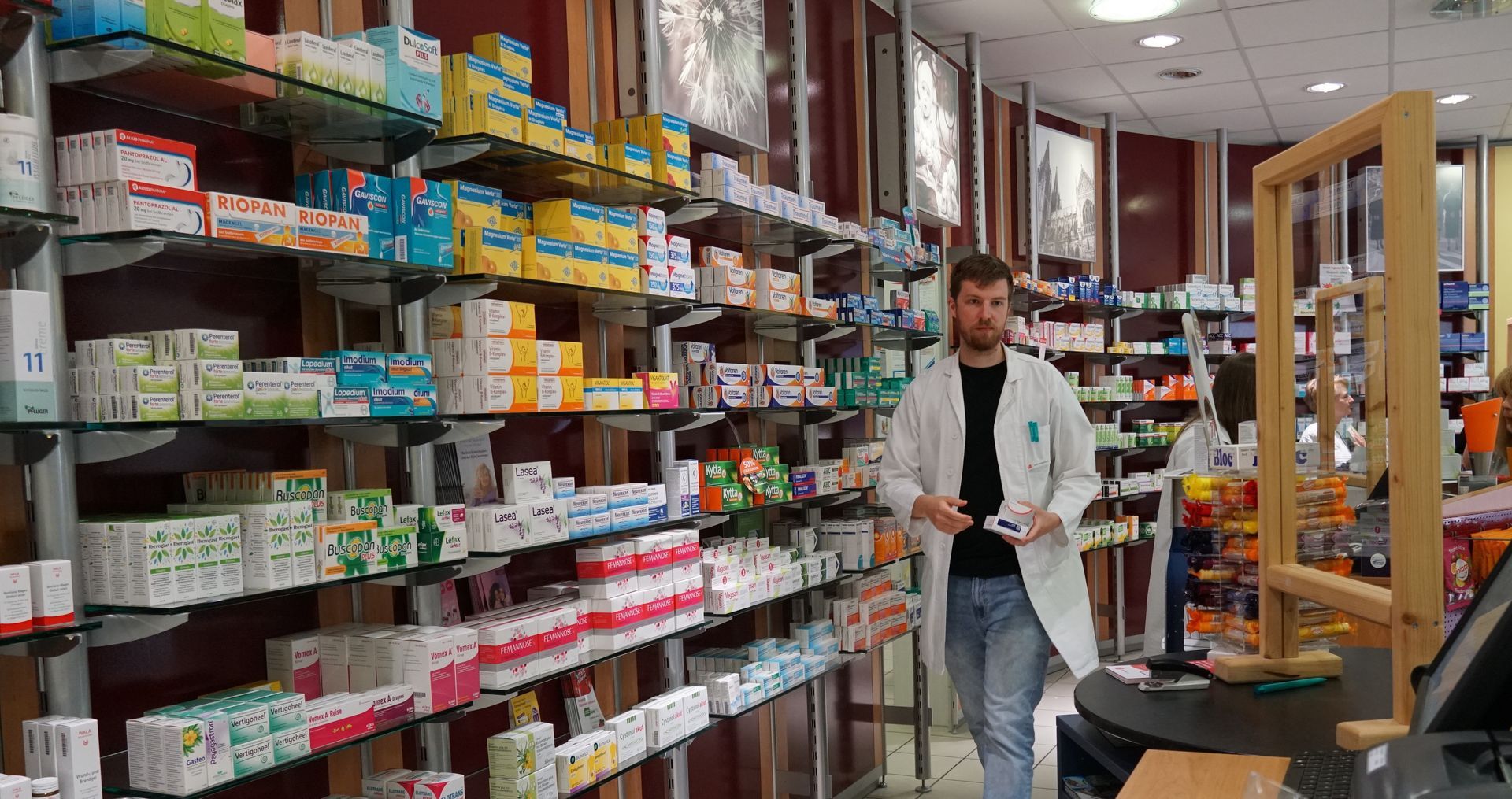 Ein Mann im weißen Kittel steht in einem Apothekenregal zwischen Medikamenten. Im Hintergrund ist eine weitere Person zu sehen.