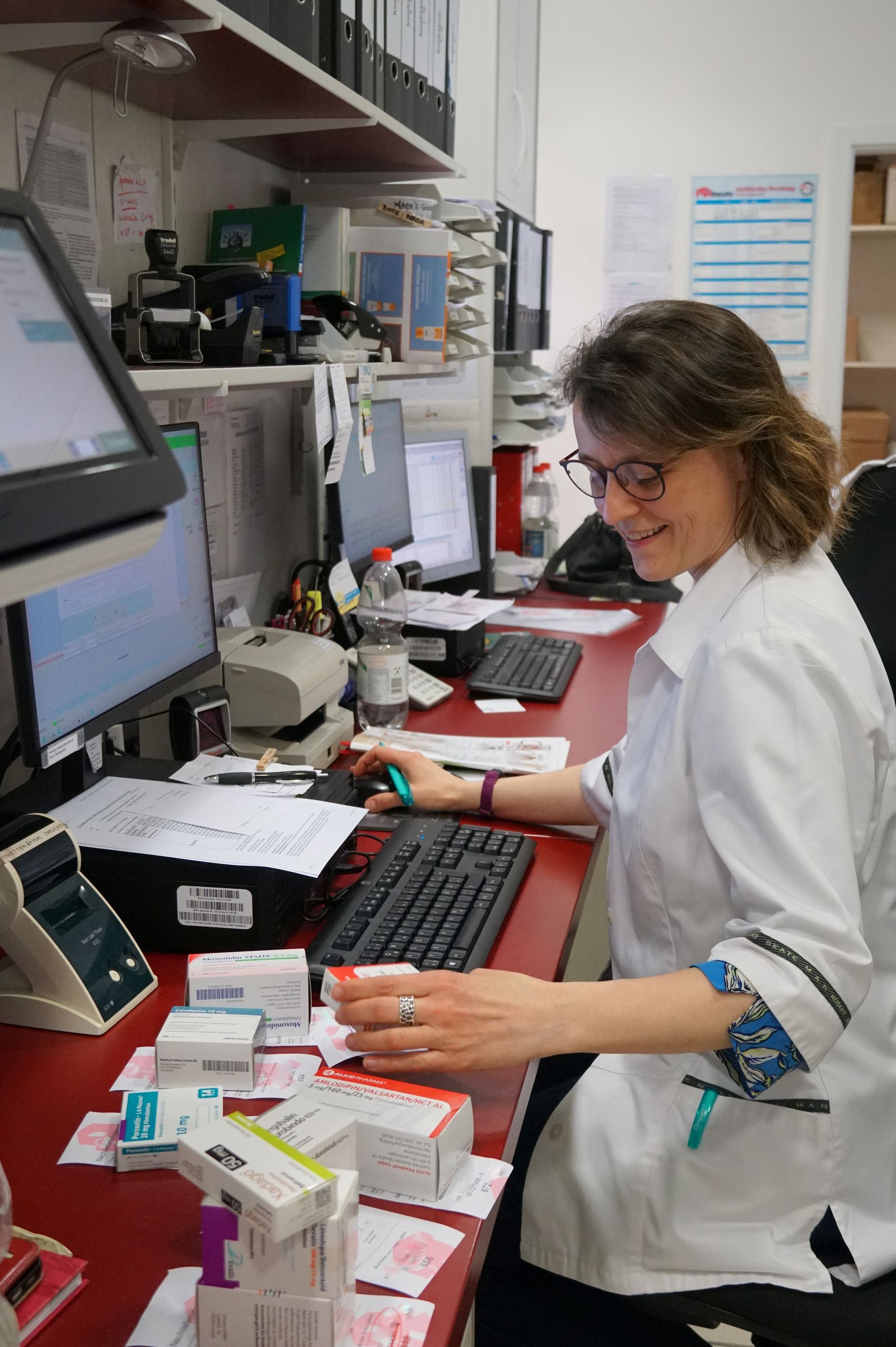 Apotheker am Schreibtisch, prüft Medikamentenpackungen. Roter Tresen, Computer, weißer Kittel, lächelnd.