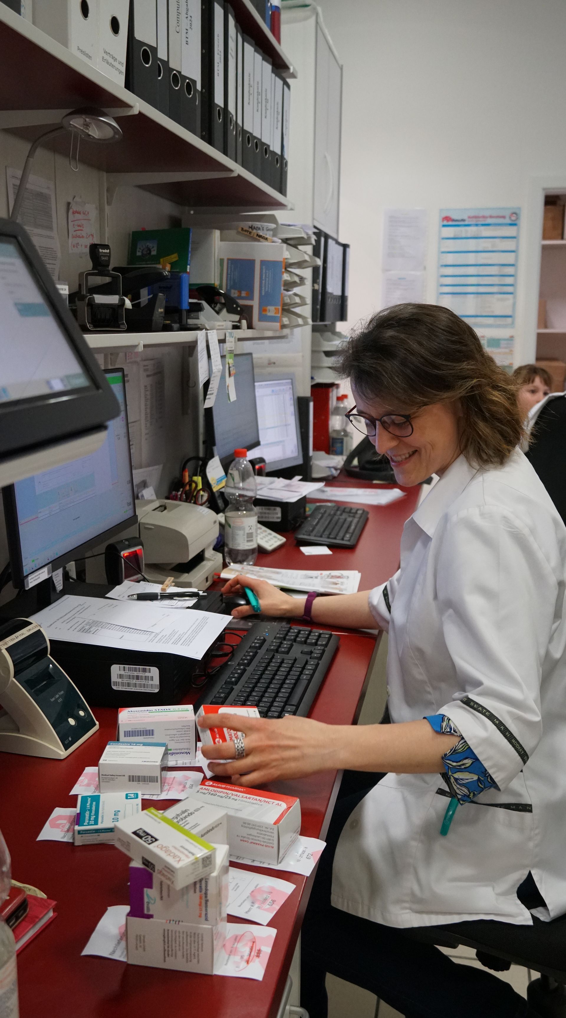 Eine Person im Laborkittel sortiert lächelnd Medikamente an einem Apothekenschalter. Schreibtisch mit Computern und Kartons.