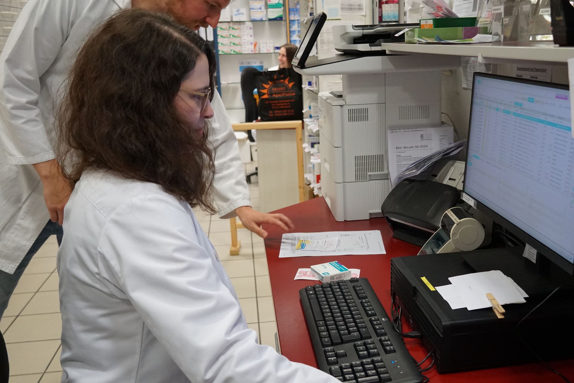 Zwei Personen in Laborkitteln an einem Apothekencomputer; eine tippt, die andere zeigt mit dem Finger.