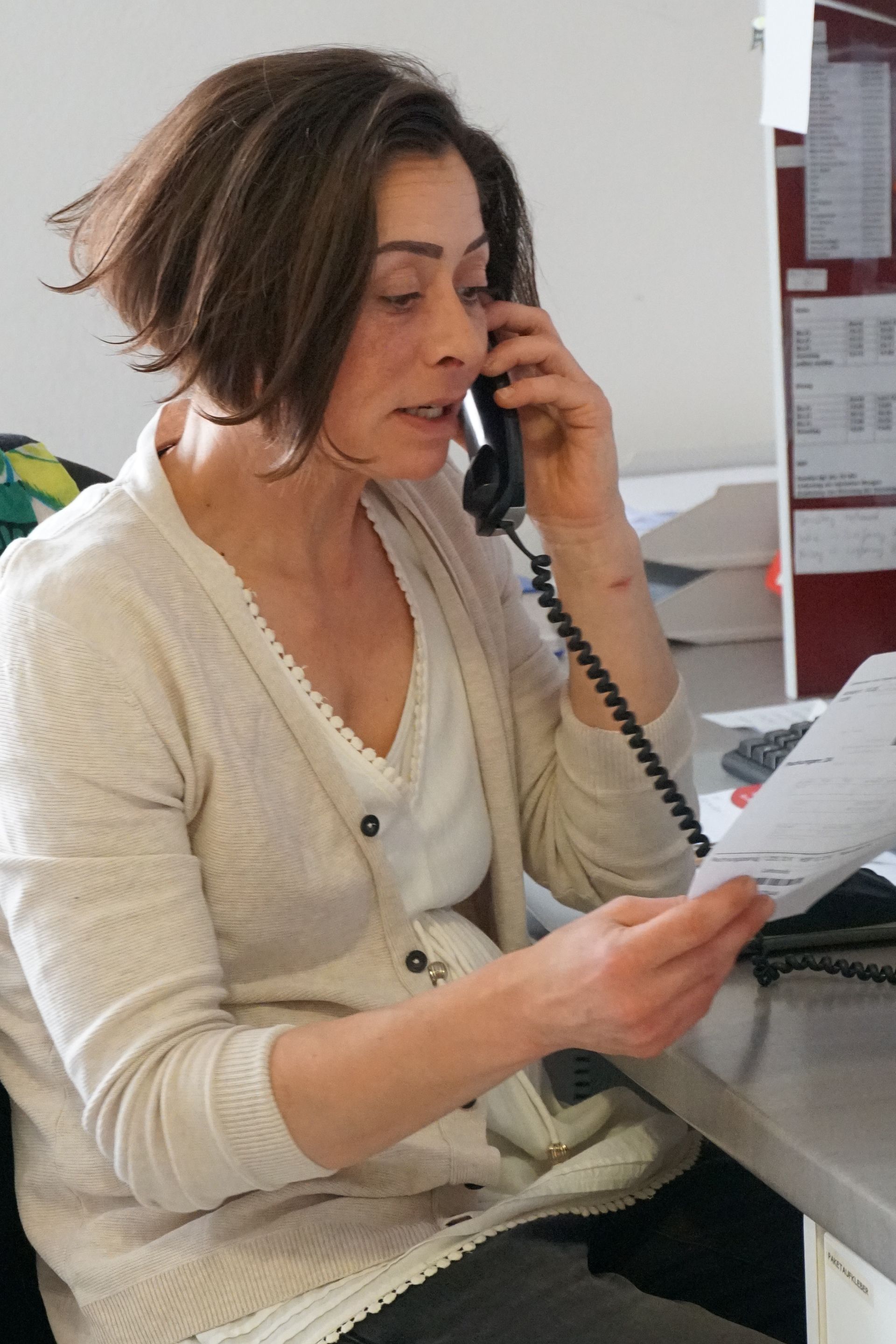 Frau mit kurzen braunen Haaren telefoniert, schaut auf einem Schreibtisch auf Papier und trägt eine weiße Strickjacke.
