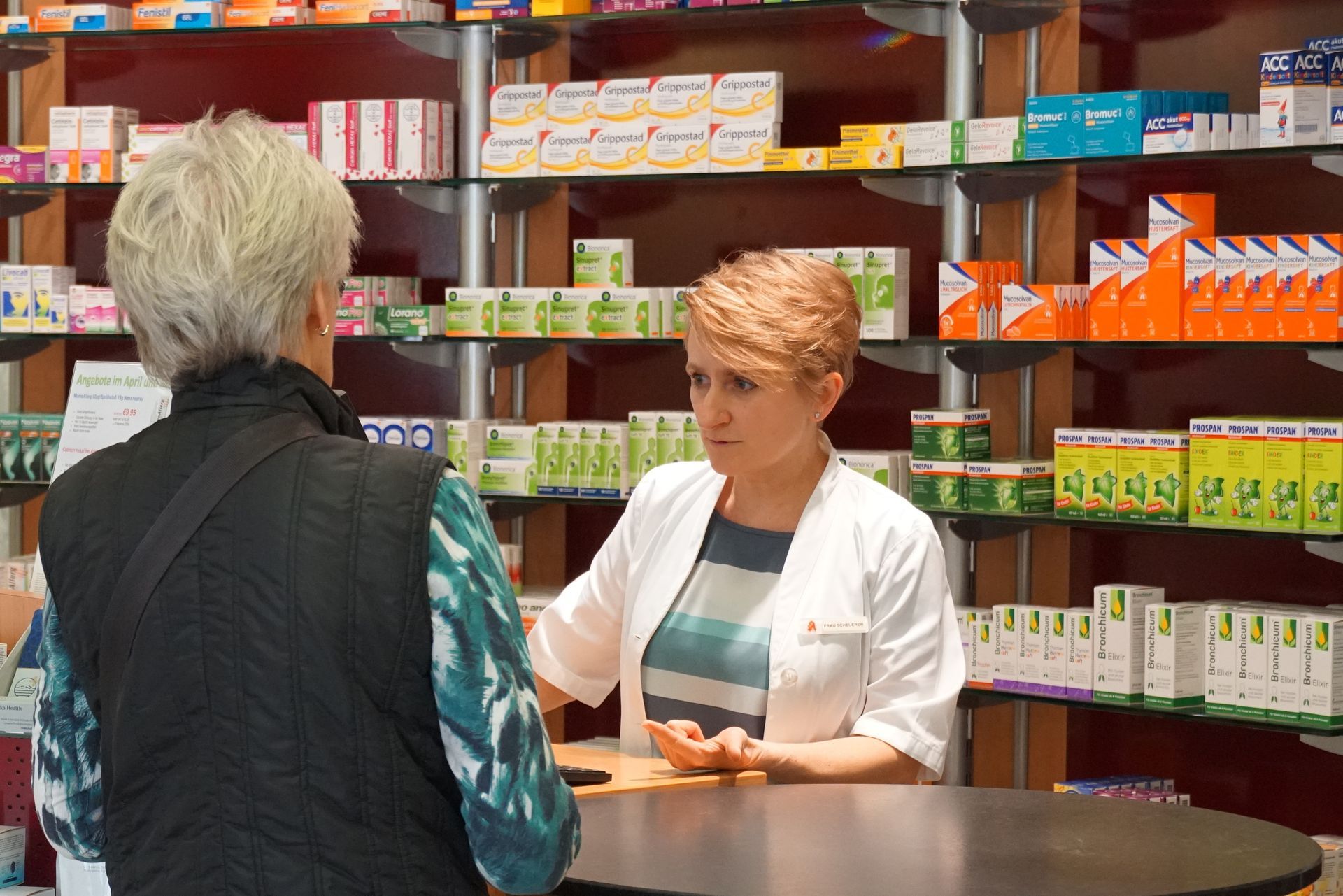 Ein Apotheker bedient einen Kunden an einem Apothekenschalter, umgeben von Medikamentenregalen.