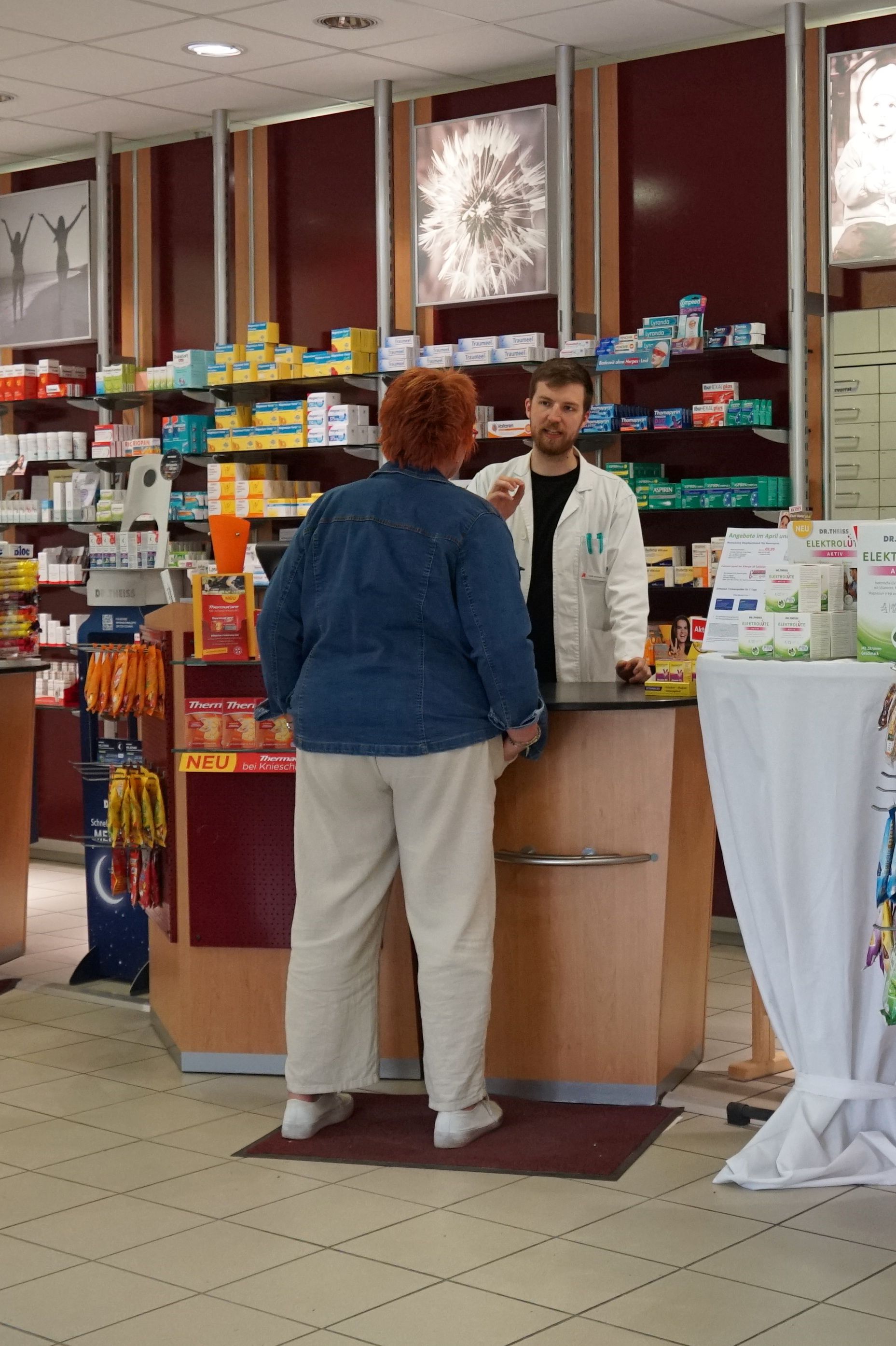 Eine Frau spricht am Apothekenschalter mit dem Apotheker. Im Hintergrund sind Regale mit Produkten zu sehen.