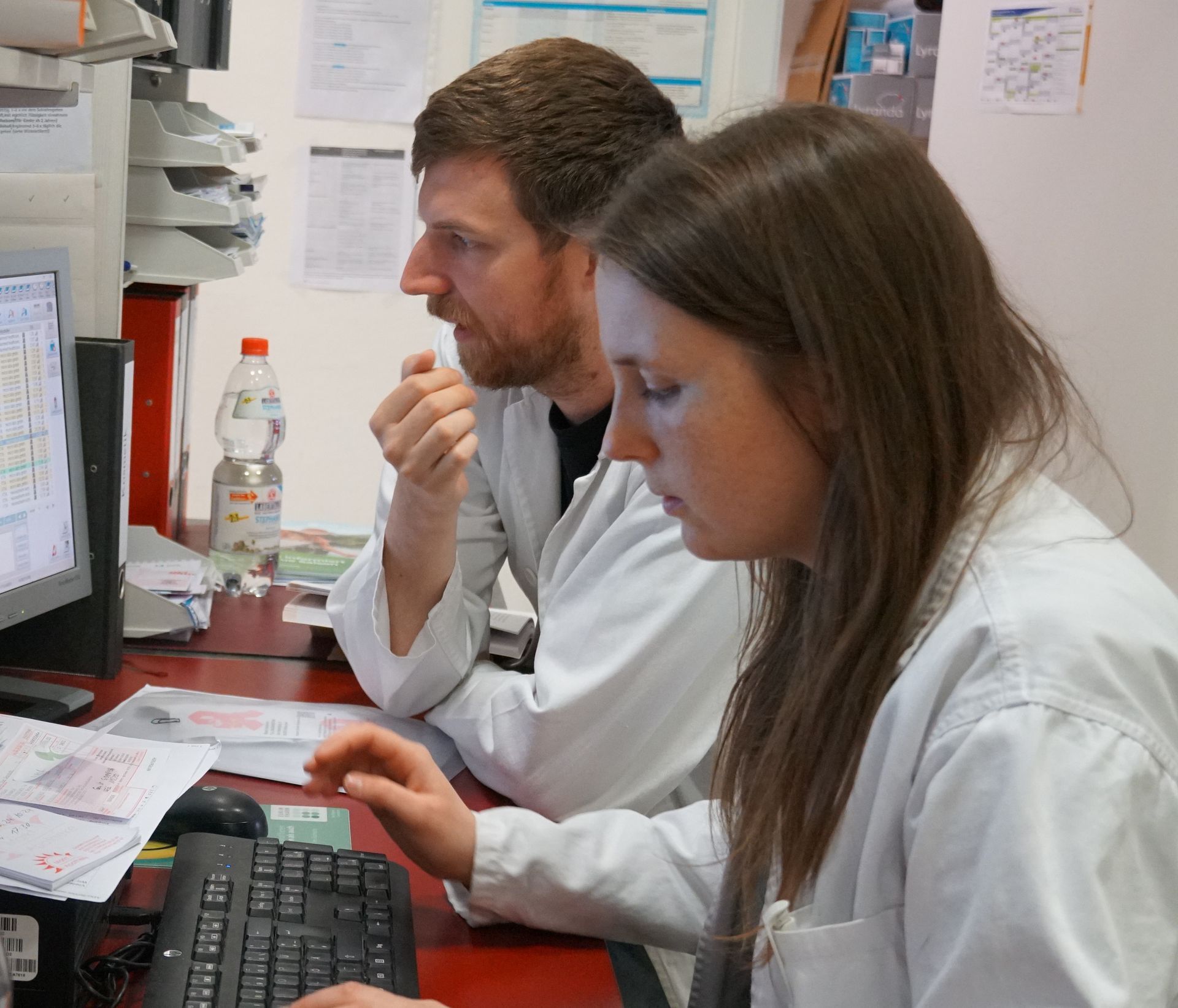 Zwei Personen in Laborkitteln schauen in einem Büro auf einen Computerbildschirm. Eine tippt.