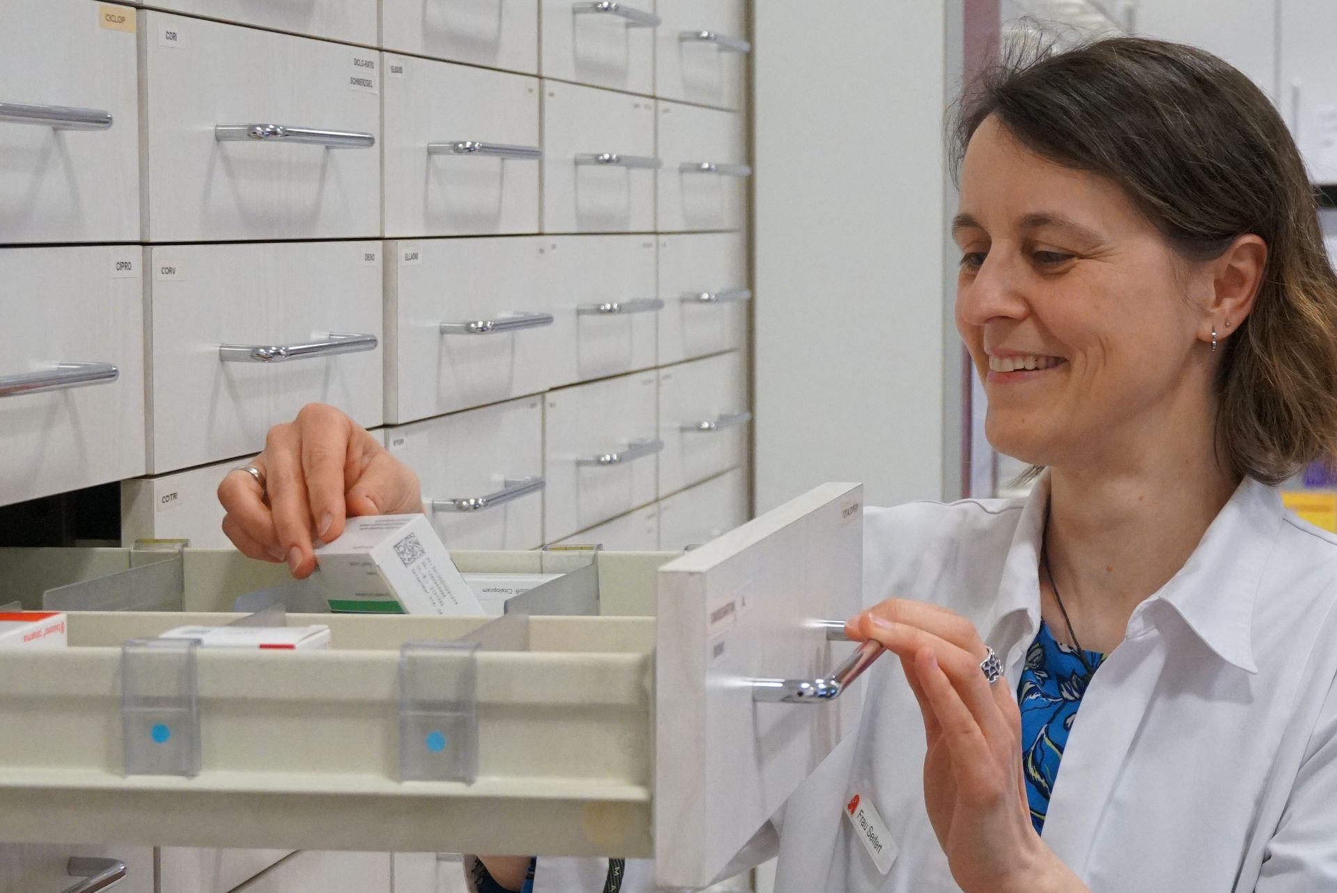 Ein Apotheker im weißen Kittel lächelt und sucht in einer Schublade des Apothekenschranks nach Medikamenten.