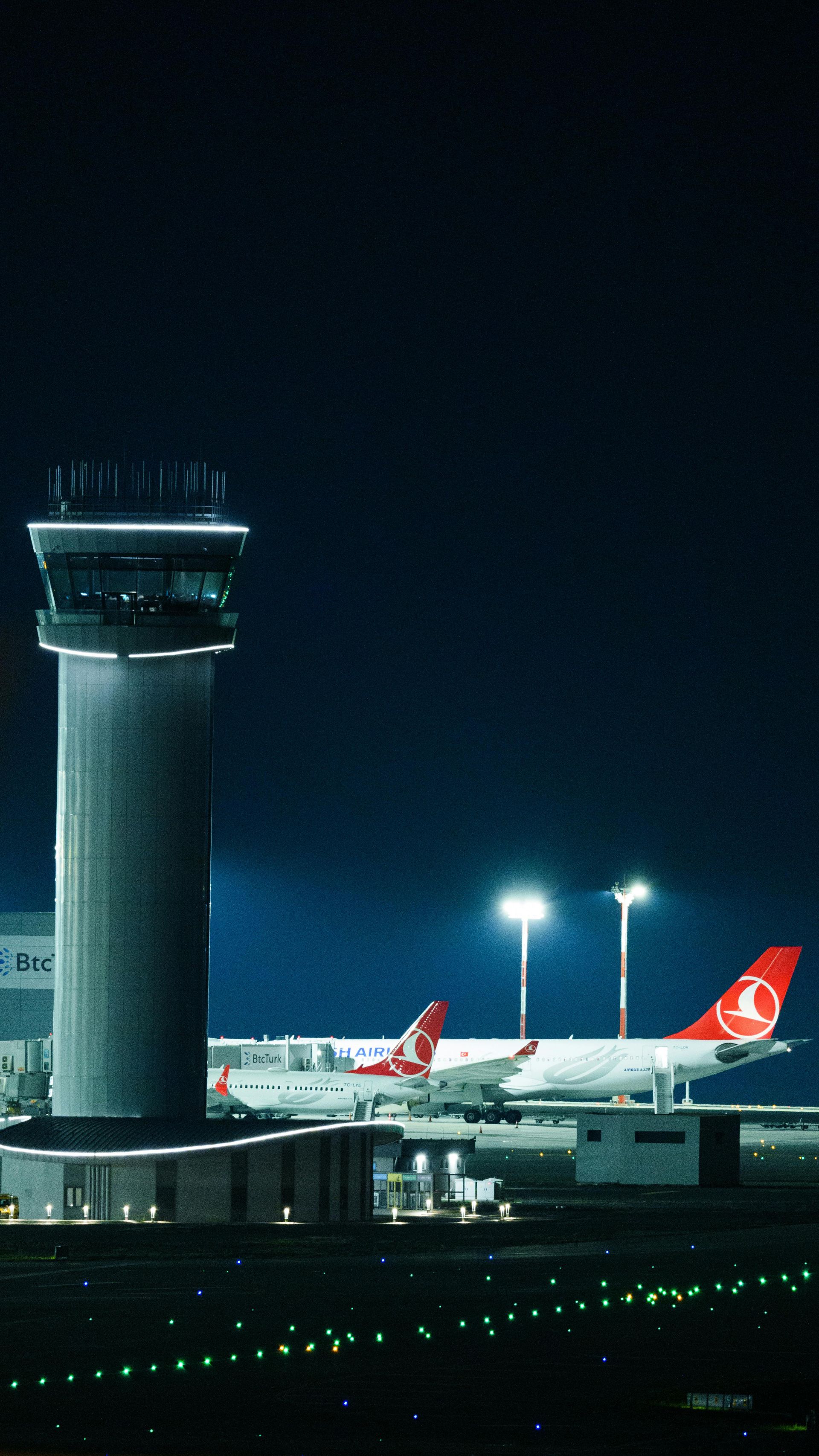 Tour de contrôle de l'aéroport située à côté d'un avion de Turkish Airlines la nuit, éclairée par les feux de piste.