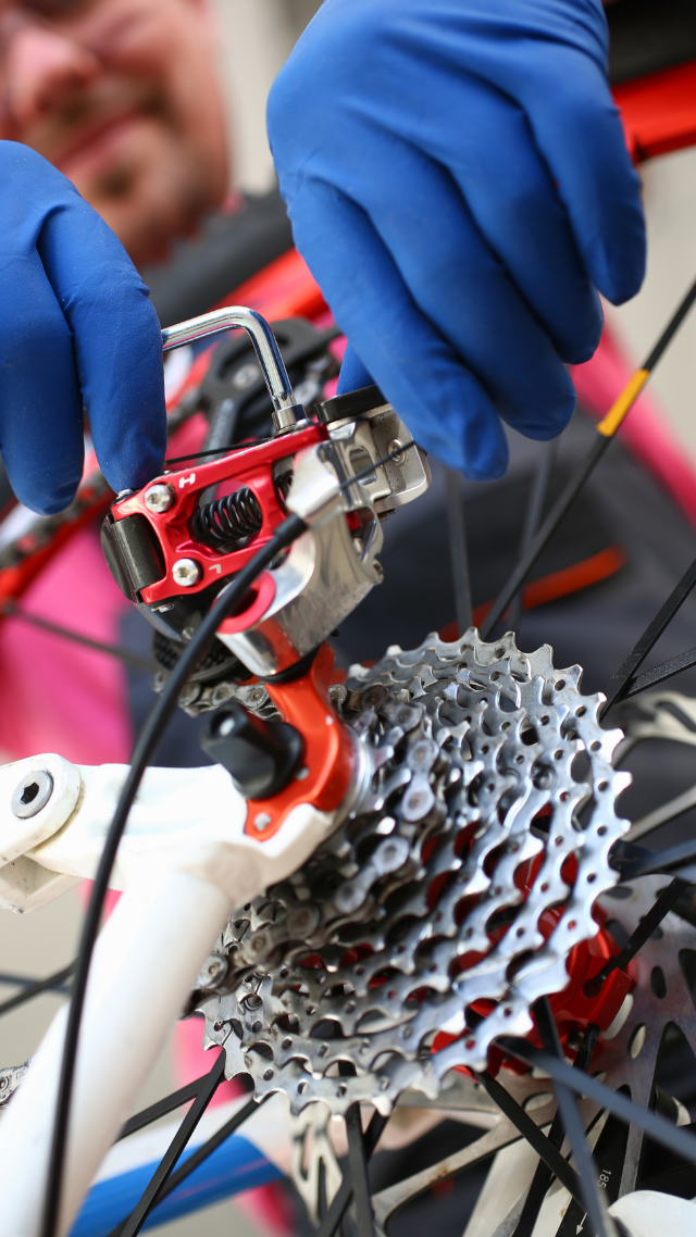 Une personne portant des gants bleus répare les engrenages d'un vélo, les composants rouges et la cassette argentée.