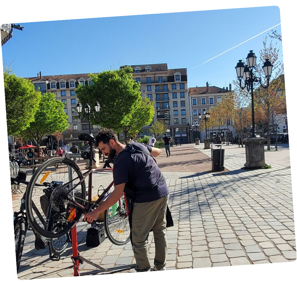 Un homme répare un vélo sur une place pavée, avec des arbres, des bâtiments et des lampadaires en arrière-plan. Ciel bleu.