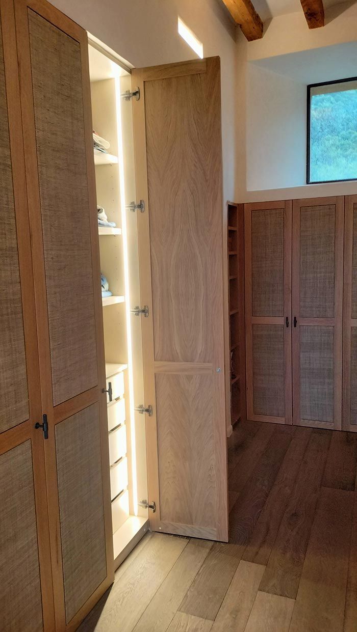 Armario de madera con puerta abierta que revela un interior iluminado, rodeado de armarios adicionales con paneles de puerta tejidos y piso de madera.