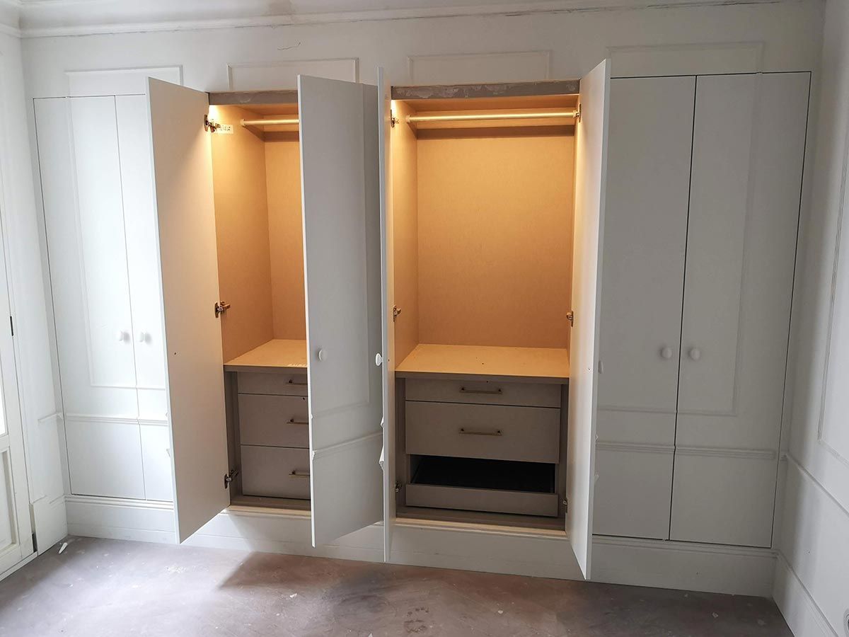 Armario empotrado blanco con puertas abiertas que revelan cajones y espacio para colgar, iluminado por luces interiores.