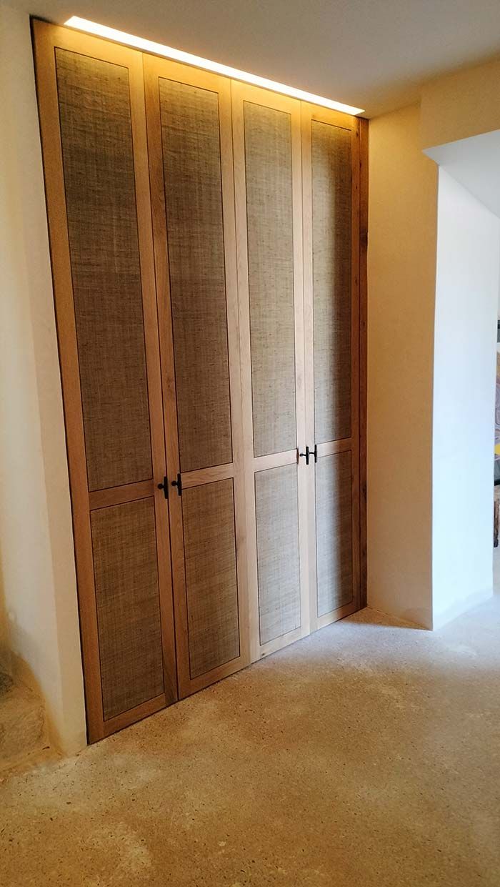 Puertas dobles de madera con paneles texturizados, manijas negras e iluminación cálida en la parte superior, ubicadas en un pasillo de tonos neutros.