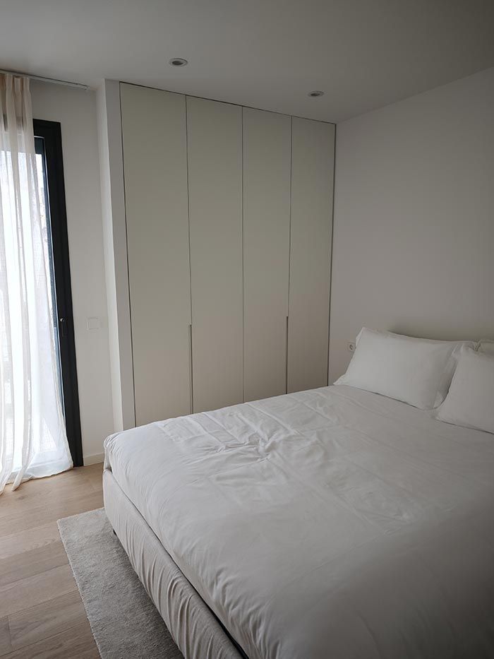 Dormitorio con cama blanca, armario y cortinas. La suave luz del sol entra en la habitación.