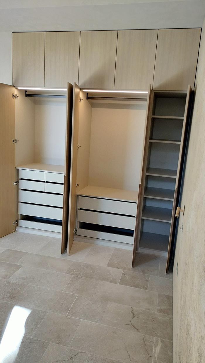 Armario empotrado con puertas, cajones, estantes y barras de madera clara. El armario está instalado sobre azulejos de color claro.