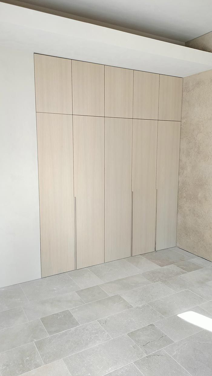 Puertas de armario con paneles de madera clara y detalles plateados, ubicadas en una habitación con piso de baldosas grises y paredes de color beige.