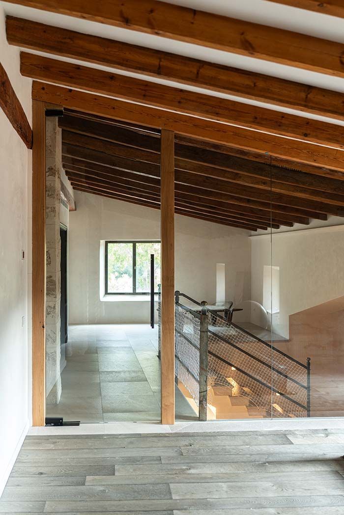 Vista interior de un edificio con vigas de madera, escalera y luz natural entrando por una ventana.