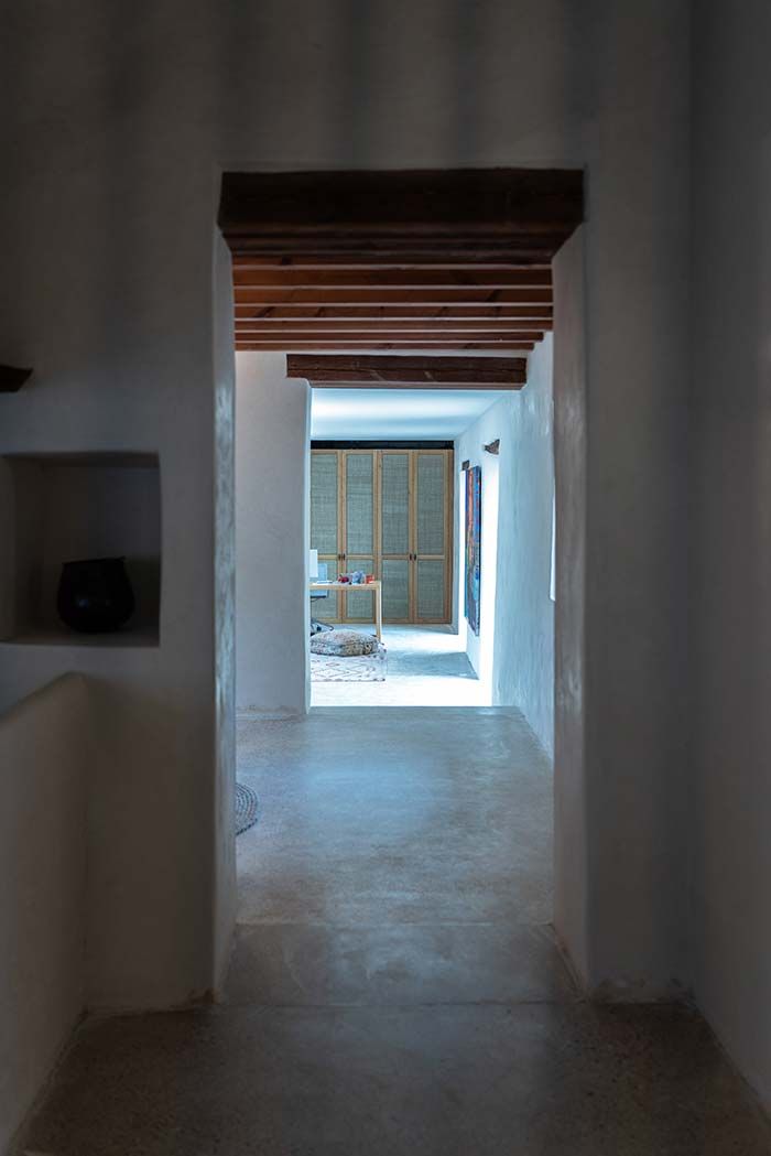 Pasillo interior con paredes blancas y vigas de madera, que da paso a una estancia más luminosa.