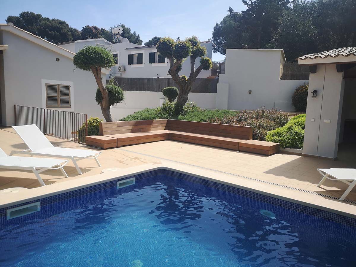 Área de piscina con agua azul, asientos y árboles cerca de un edificio blanco.