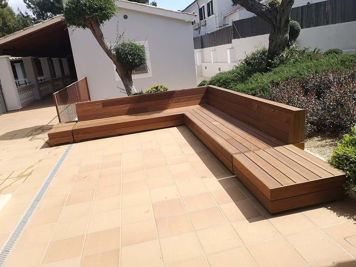 Banco de madera en forma de L en un patio de baldosas. Luz solar y arbustos.