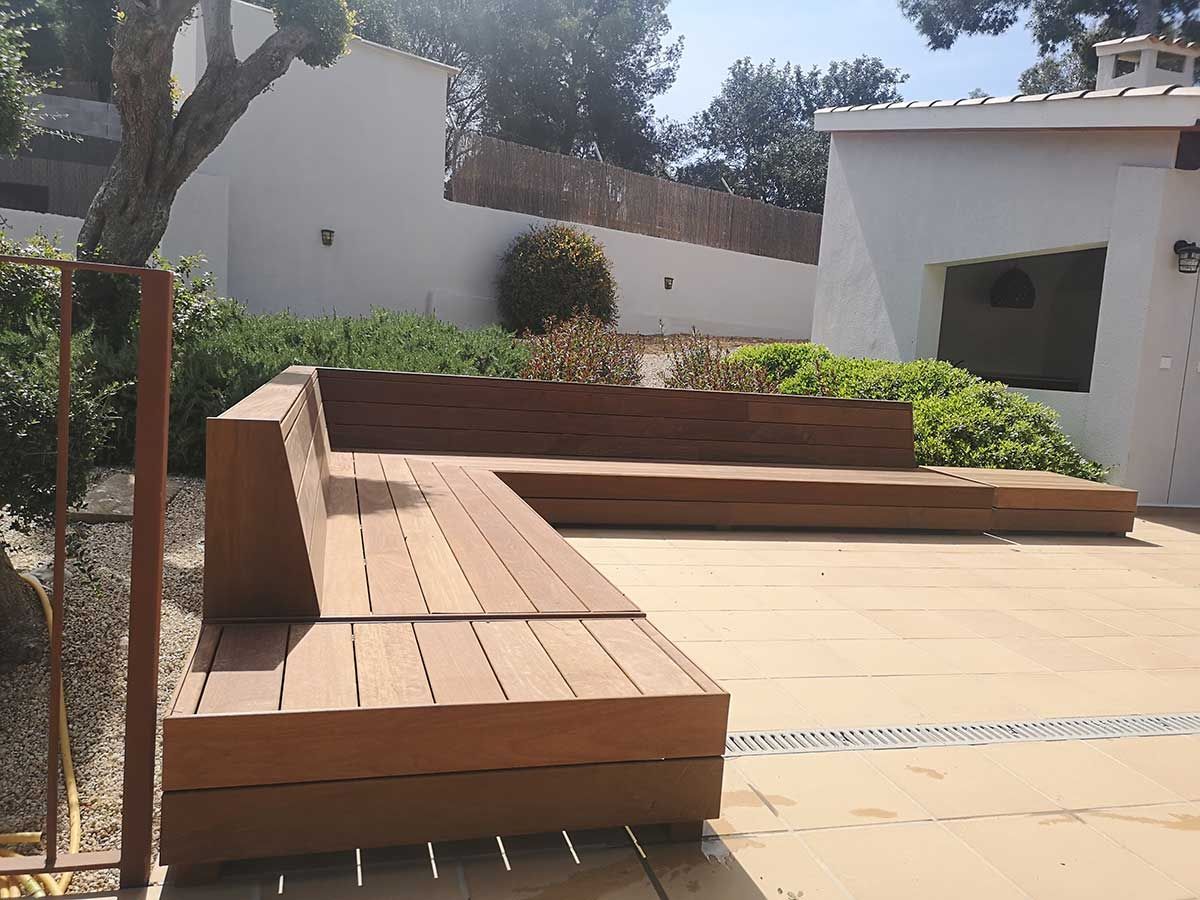 Zona de estar al aire libre de madera en un patio, con un edificio blanco y vegetación al fondo.