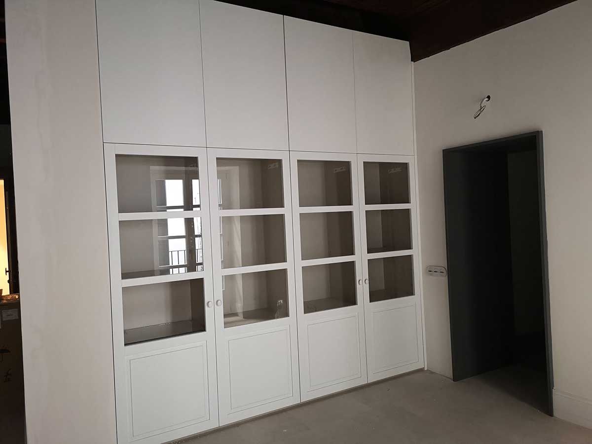 Unidad de almacenamiento empotrada de color blanco con puertas con paneles de vidrio y gabinetes superiores de color blanco sólido en una habitación con puerta.