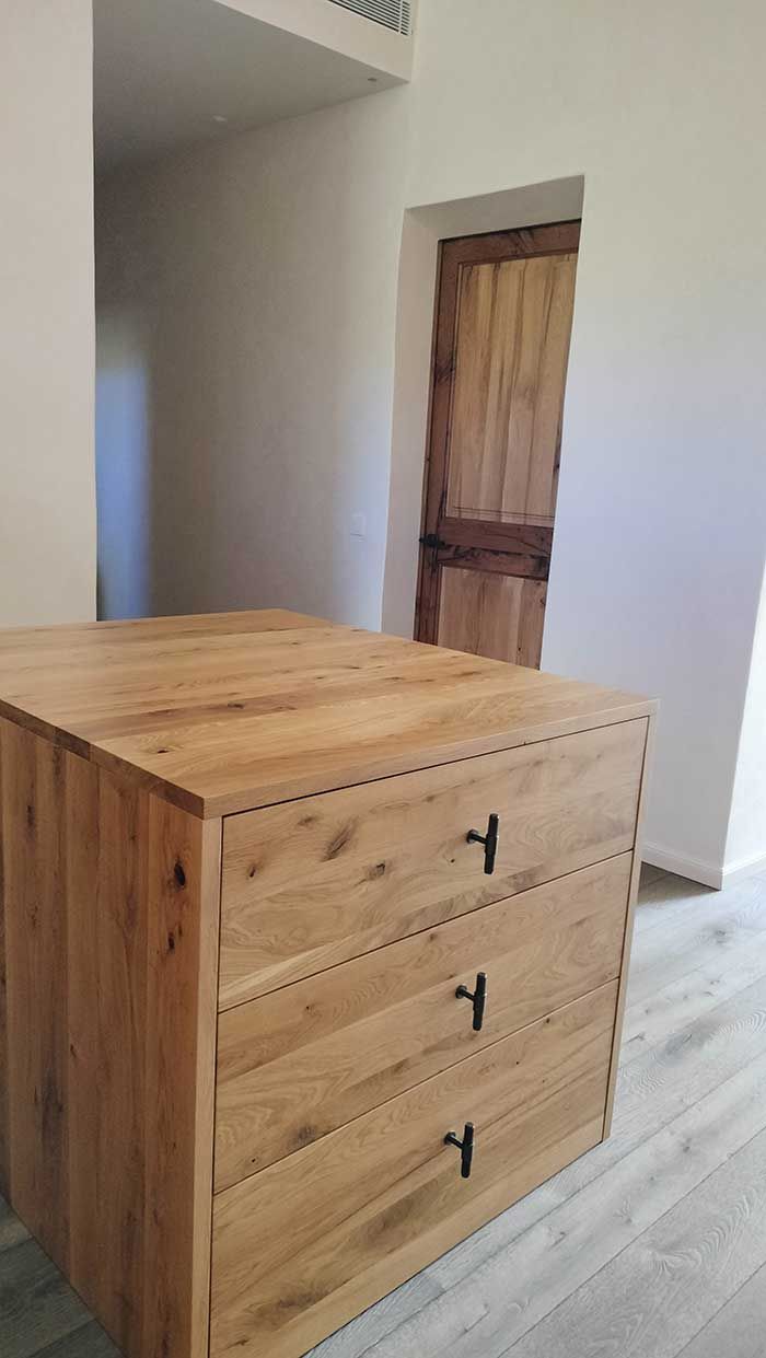 Cómoda de madera con tres cajones, en una habitación con puerta de madera y paredes blancas.