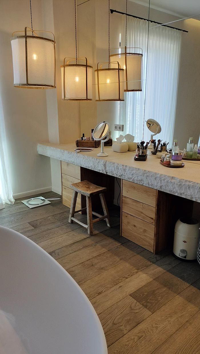 Baño con encimera de color claro y tocador con paneles de madera. Se ven taburete, grifería de lavabo y lámparas colgantes.