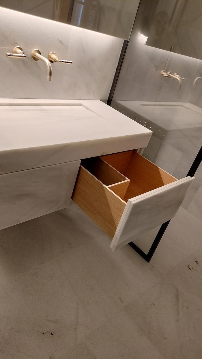 Mueble de baño con cajón abierto, apariencia de mármol gris, accesorios dorados e interior de cajón de madera.