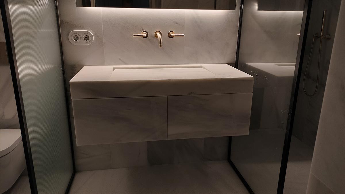 Moderno baño de mármol con tocador flotante, grifo dorado y mampara de ducha de vidrio.