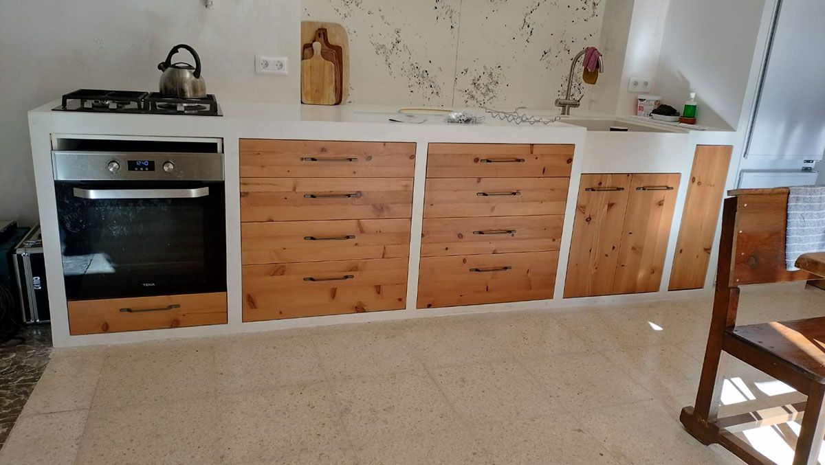 Cocina con muebles de madera y horno. Hornilla, tetera y vajilla sobre encimera blanca.