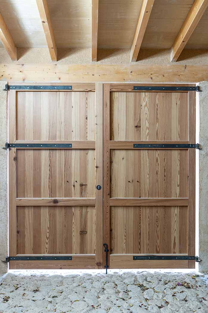 Puertas dobles de madera con bisagras y pestillo de metal negro, colocadas en una pared de piedra debajo de vigas de madera.