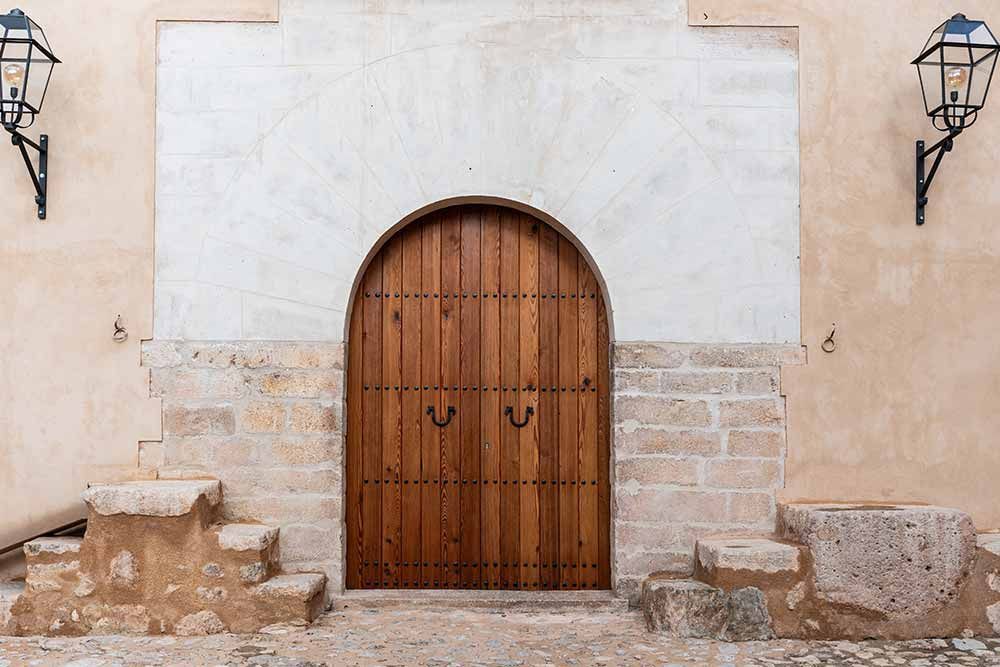 Puerta arqueada de madera empotrada en pared de piedra y estuco, con faroles a ambos lados.