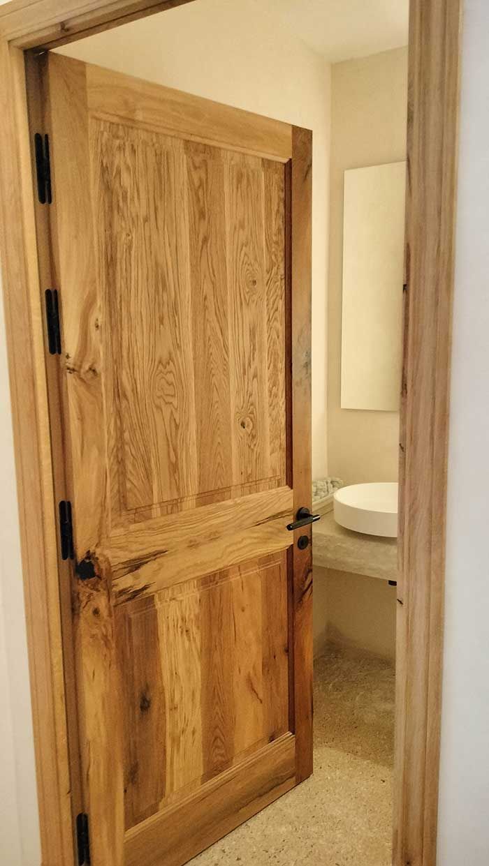 Puerta de madera entreabierta, revelando un baño con lavabo, espejo y suelo de color claro.