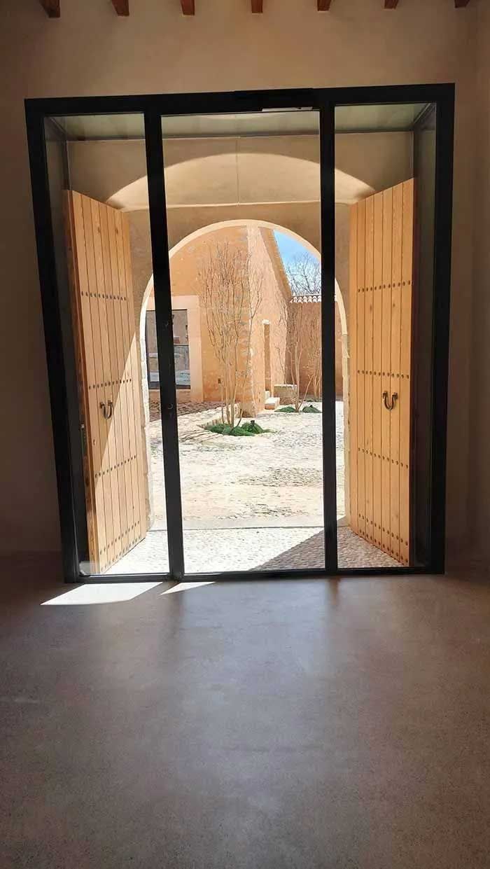 Unas puertas de madera dan a un patio de piedra. La luz del sol entra a raudales por la puerta.