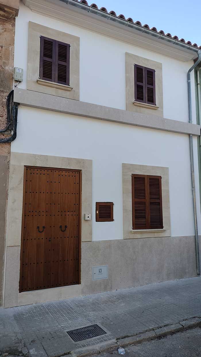 Edificio blanco con persianas y puerta marrones, situado en una calle ligeramente inclinada.