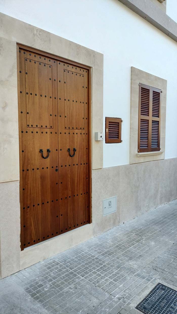 Puerta de madera con herrajes ornamentados y una ventana con contraventanas en una fachada de edificio de color claro.