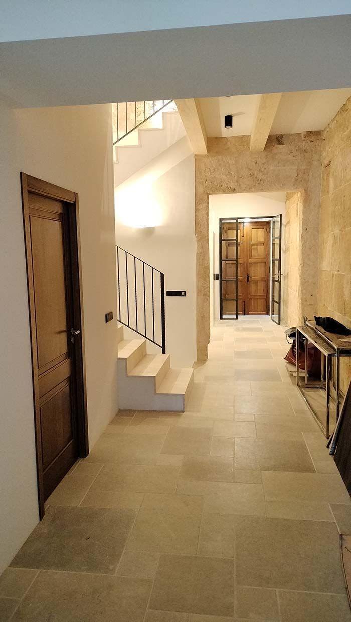 Pasillo con suelo de piedra, escalera con barandilla blanca y negra, puerta de madera y paredes de piedra.