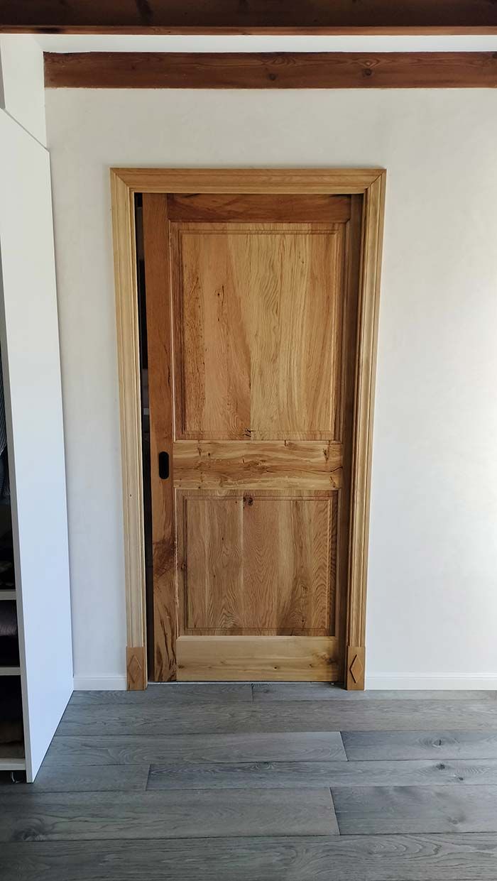 Puerta de madera con marco a juego, en pared blanca con molduras de madera. Suelo gris.